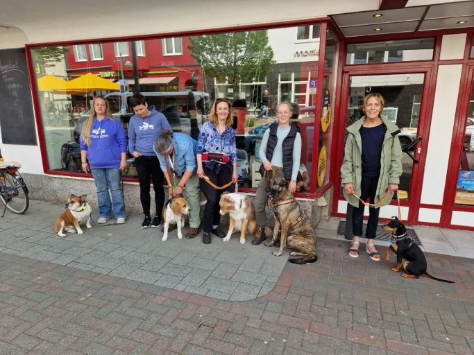 Sechs Personen mit verschiedenen Hunden stehen vor einem Geschäft.